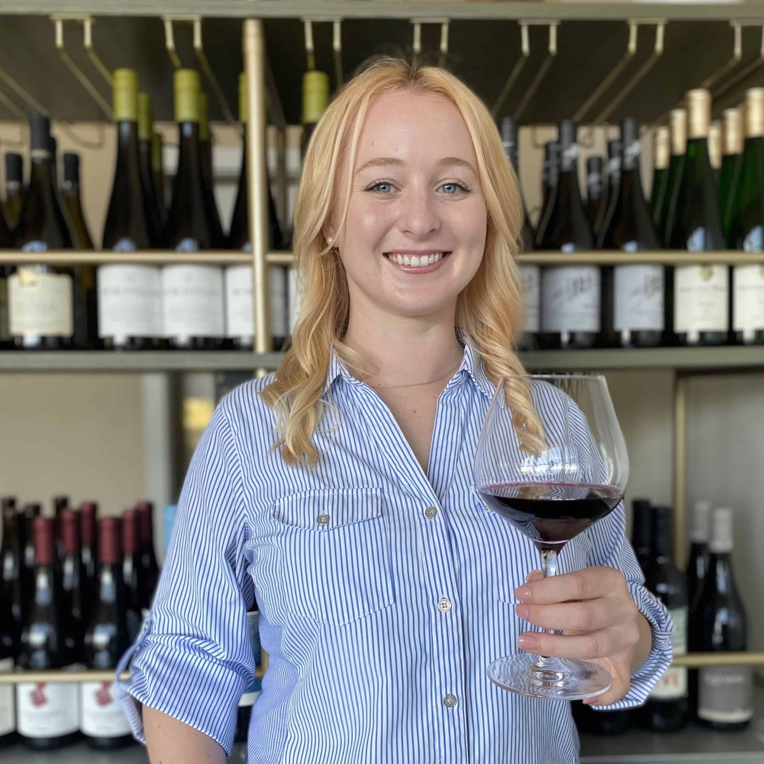 Jenna Isaacs standing in front of a wine rack and holding a glass of red wine
